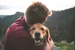 Un homme enlace affectueusement un chien heureux, avec un paysage naturel en arrière-plan.