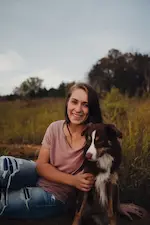Une femme assise dans un champ sourit à côté de son chien, un berger australien.
